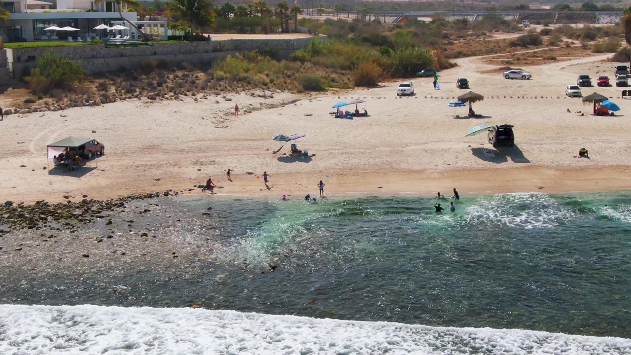 At El Tule Beach in Los Cabos, a drone view shows several people near the waterline, clear surf over rocks, umbrellas on pale sand, parked vehicles, and low vegetation behind the beach.