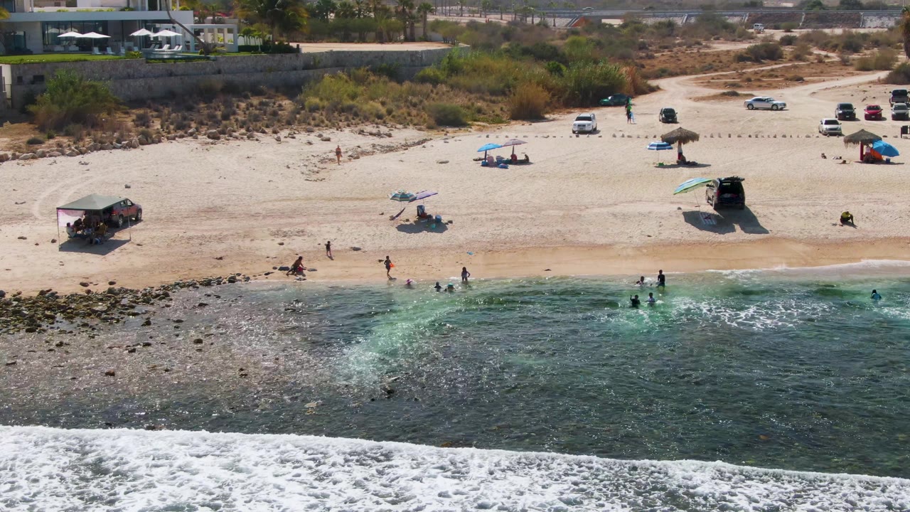 Drone view of beachgoers by clear water with umbrellas, parked cars, and surf.