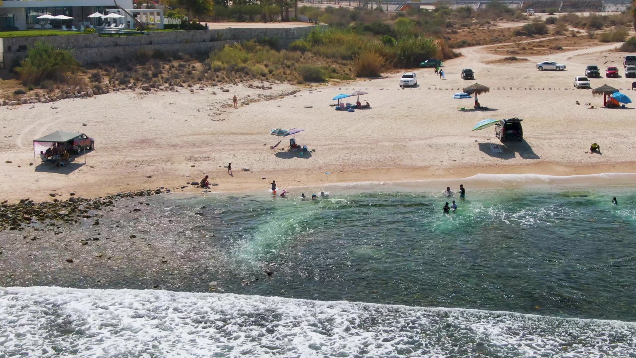 At El Tule Beach in Los Cabos, a foamy wave reaches farther across the foreground as small groups of people stand near the water and beach umbrellas and vehicles remain fixed on the sand.