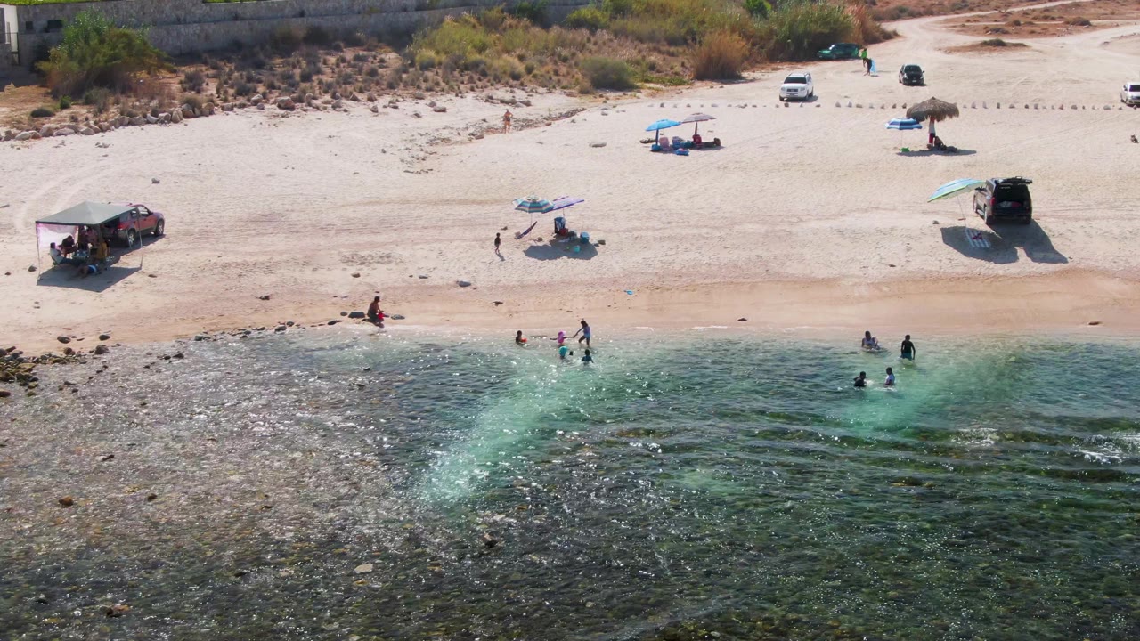 Aerial view at El Tule Beach in Los Cabos showing swimmers in shallow green-blue water, pale sand, shade umbrellas, and a few vehicles parked along the beach.