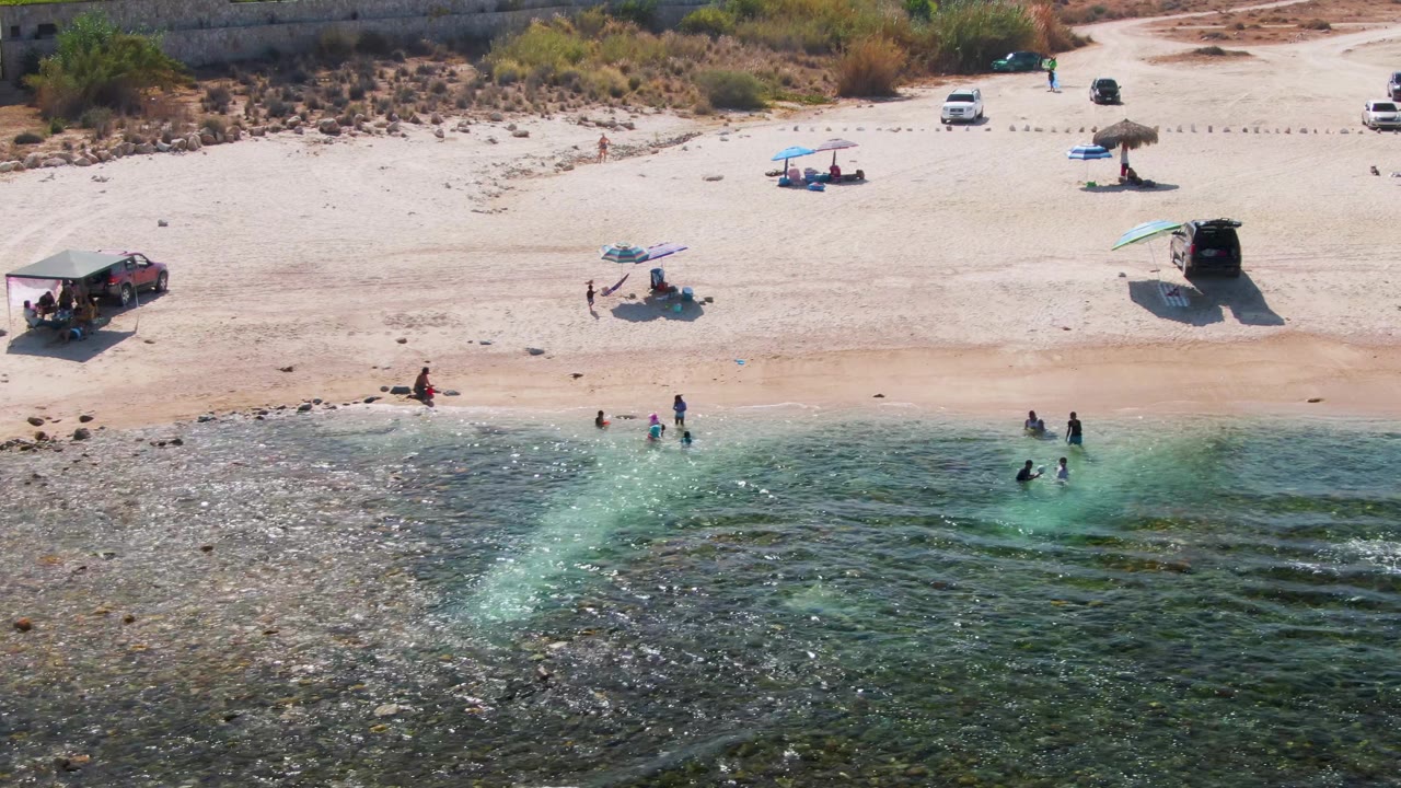 Beachgoers wade in clear water beside a sandy beach with scattered umbrellas and vehicles.