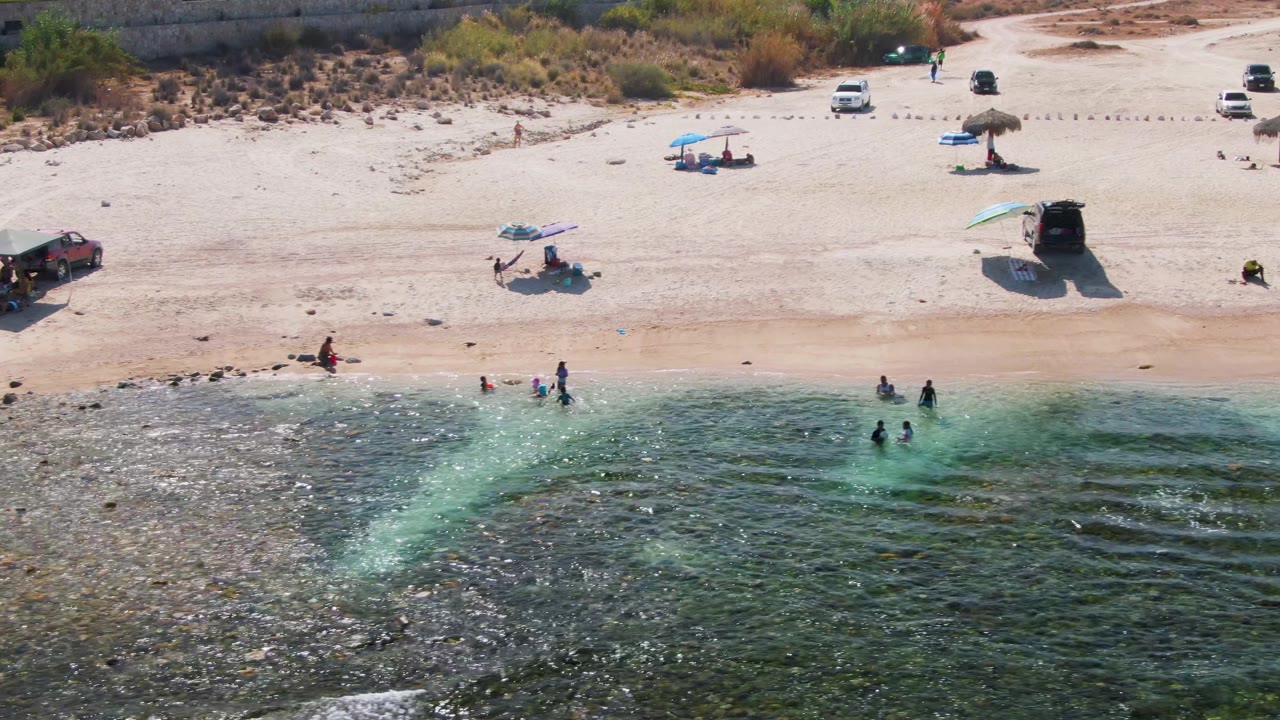 El Tule Beach appears from above with rippled green water in the foreground, beachgoers near the surf line, a straw-style umbrella, a blue umbrella, and vehicles on the sand.