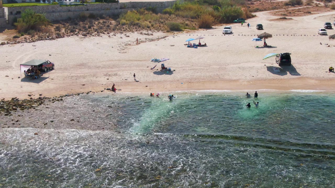 Aerial frame at El Tule Beach in Los Cabos showing swimmers near the shoreline, umbrellas on the sand, parked vehicles above the beach, and rocky green-blue water in the foreground.