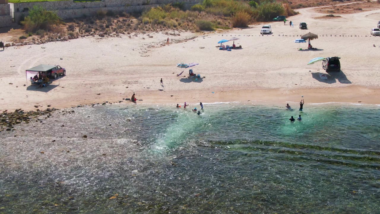El Tule Beach aerial frame showing beachgoers spread between the water and sand, darker rocky surf in the foreground, and umbrellas and parked vehicles set back from the shore.