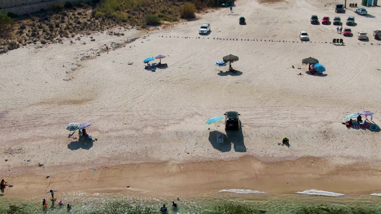 Aerial view of El Tule Beach in Los Cabos with scattered umbrellas and parked cars on pale sand, people wading near the shoreline, and green water along the lower edge.