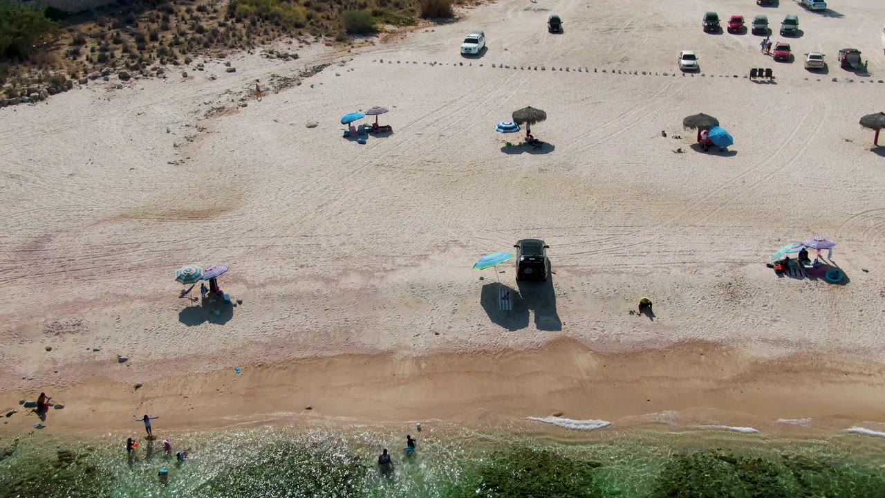 Drone view of sandy beach with umbrellas, vehicles, and people in shallow surf.