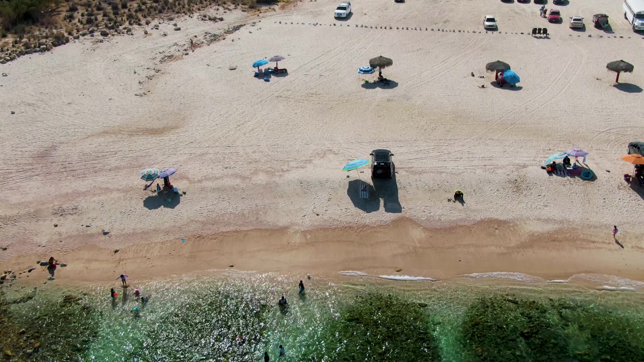 Overhead view at El Tule Beach with sun glare on green water, people spaced along the surf, umbrellas on the dry sand, and parked vehicles near the upper beach.