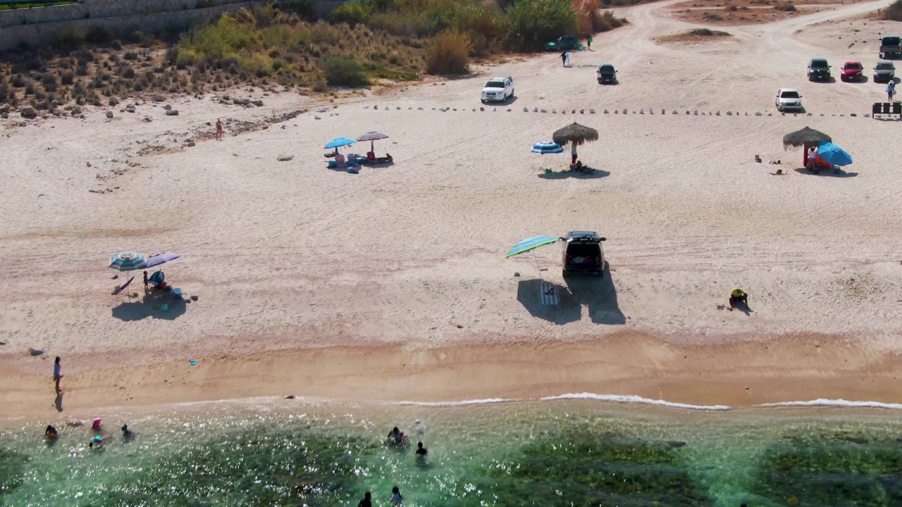 At 0:02, the drone view over El Tule Beach shows small figures wading near the lower edge, turquoise patches in the water, umbrella shade on the sand, and multiple parked cars near the dunes.
