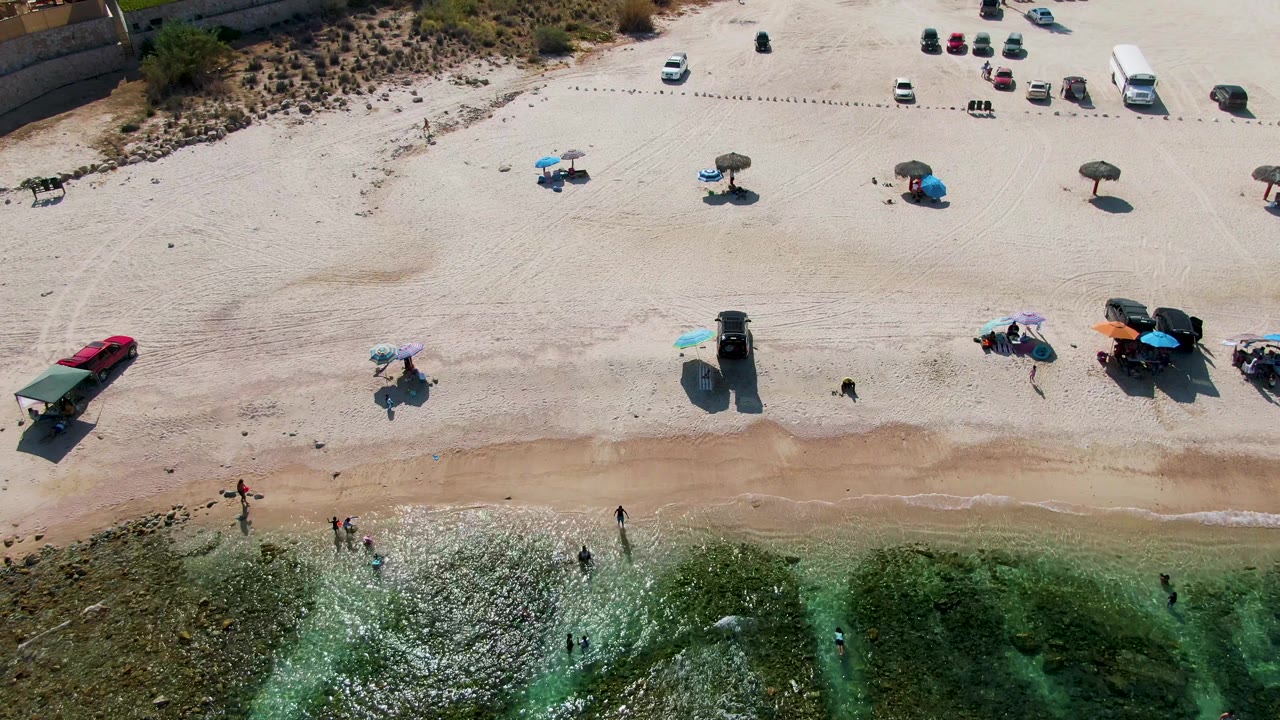 Overhead drone-style view at El Tule Beach in Los Cabos showing green shallow water, wet sand, scattered umbrellas, parked cars, and small people near the shore.