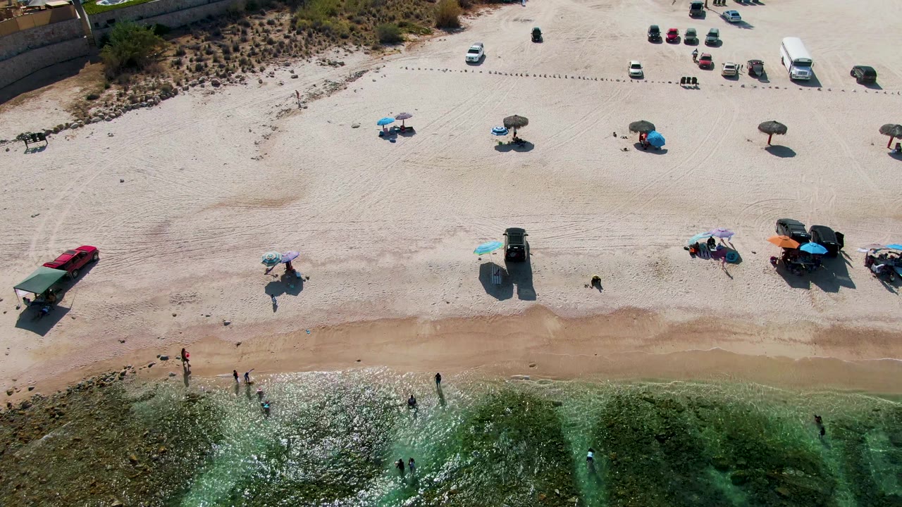 Aerial beach frame with wet shoreline, sparse umbrellas, parked cars, and people in shallow water.