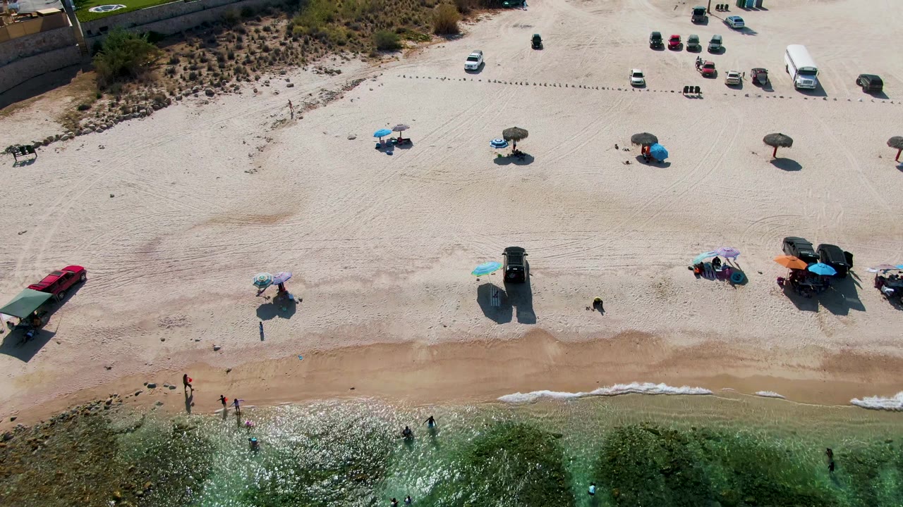At 0:02, the El Tule Beach shoreline remains centered with green water at the lower edge, wet sand exposed above it, people near the water, and parked vehicles and umbrellas farther up the beach.
