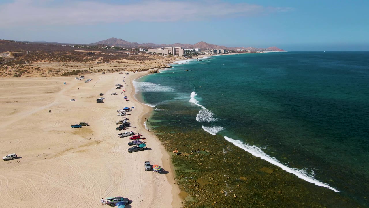 Aerial view of El Tule Beach in Los Cabos with parked vehicles on pale sand, green-blue water, white surf, distant hills, shoreline buildings, and no visible people.
