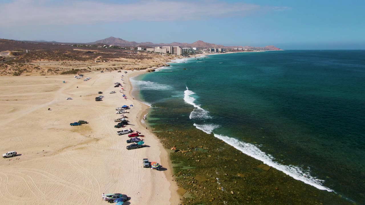 Wide drone view of beach vehicles, shade umbrellas, and waves rolling toward shore.