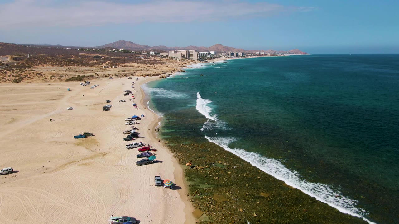El Tule Beach shoreline curves toward distant Los Cabos buildings, with parked cars on the sand, darker green shallows, white breaking waves, low hills, and no visible people.