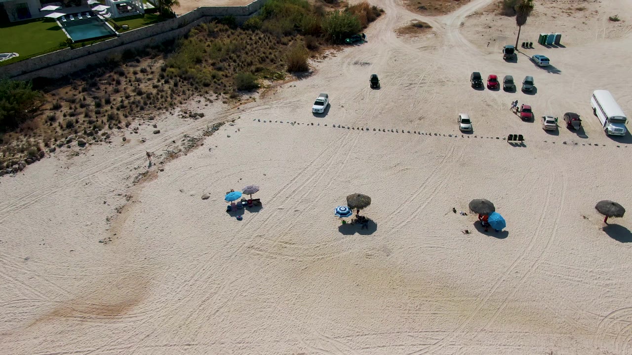 Overhead drone frame at El Tule Beach Los Cabos showing pale sand, parked vehicles, umbrellas with small beachgoers, dune scrub, and a wall along the upper edge.
