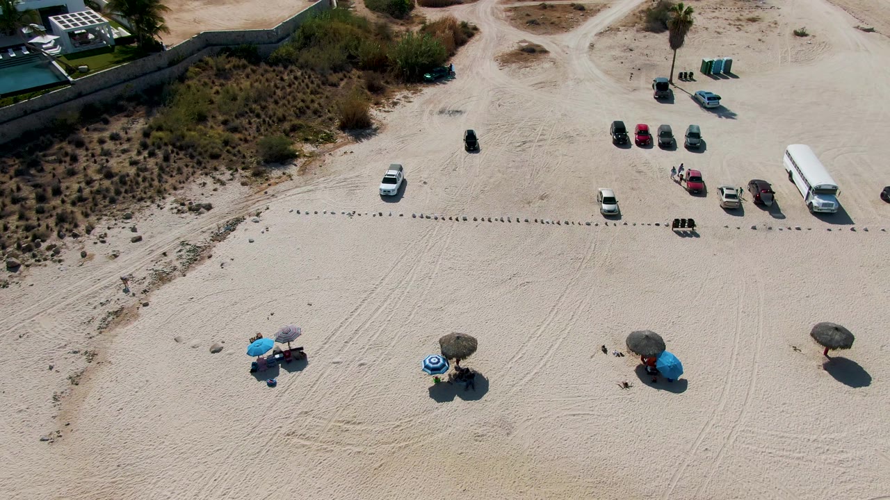 Drone frame at El Tule Beach Los Cabos showing the vehicle cluster shifted toward the right side, umbrellas spread across the lower sand, and scrubby dunes and access roads above.