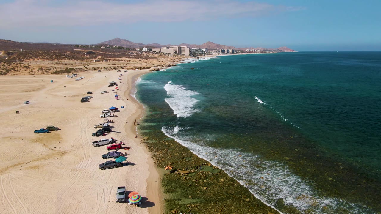 Aerial frame over El Tule Beach Los Cabos showing parked vehicles and canopies along the sand, green-blue surf breaking near shore, distant hills, and buildings on the coastline.