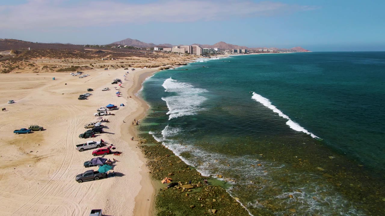 Wide drone-like frame of El Tule Beach Los Cabos with parked cars along the left side of the sand, deeper blue water to the right, white surf bands, hills, and buildings in the distance.