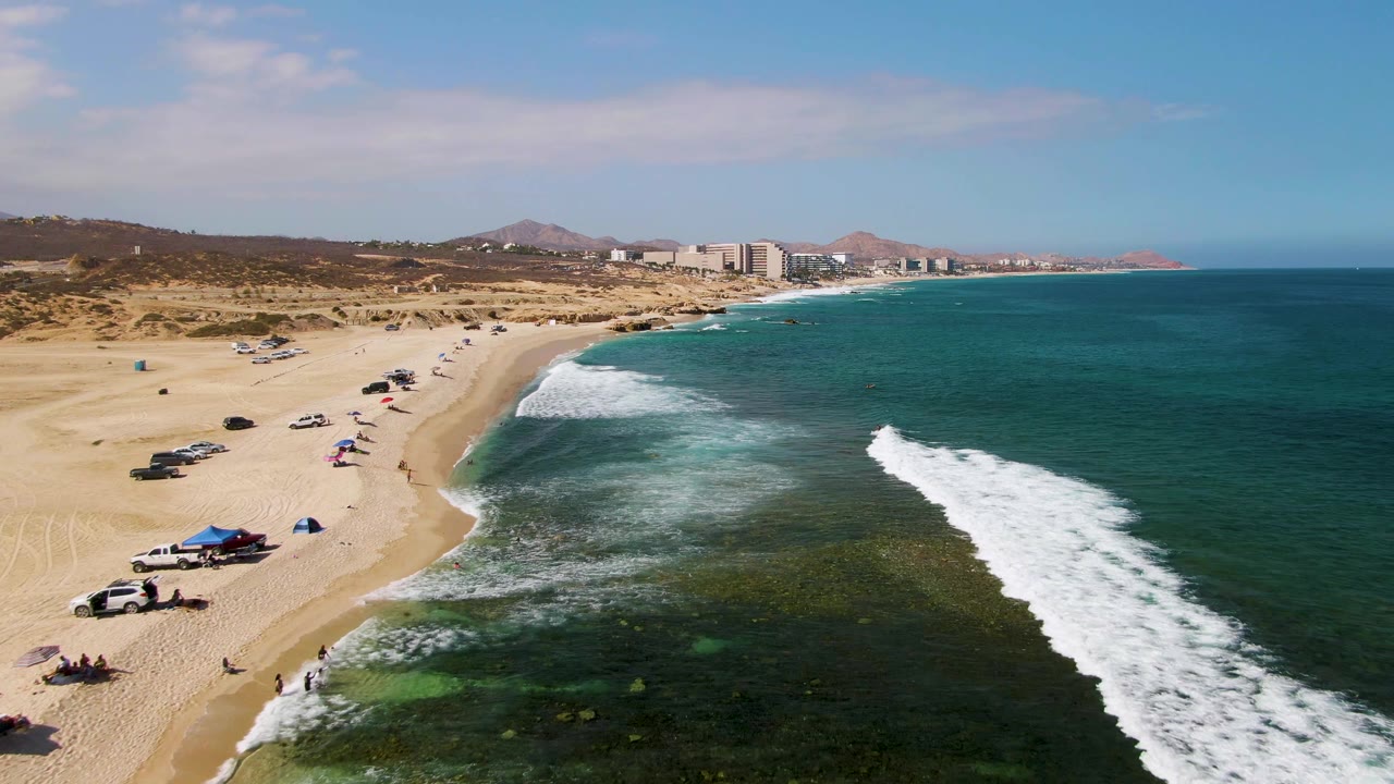 Aerial frame at El Tule Beach Los Cabos showing a sandy beach with parked cars and umbrellas on the left, clear water and white surf on the right, and distant hills and buildings beyond the coastline.