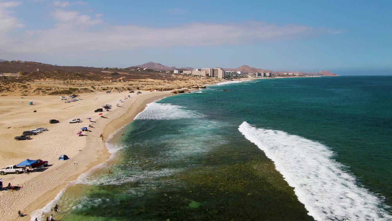 Beach shoreline with clear water, white surf, umbrellas, cars, and distant buildings.