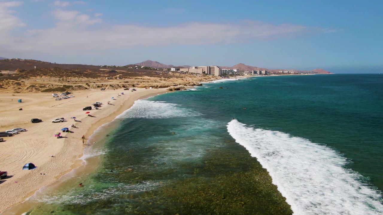 Aerial frame at El Tule Beach Los Cabos showing breaking waves over darker shallow water, a curving sandy beach with scattered vehicles and umbrellas, and the coastline receding toward distant buildings.