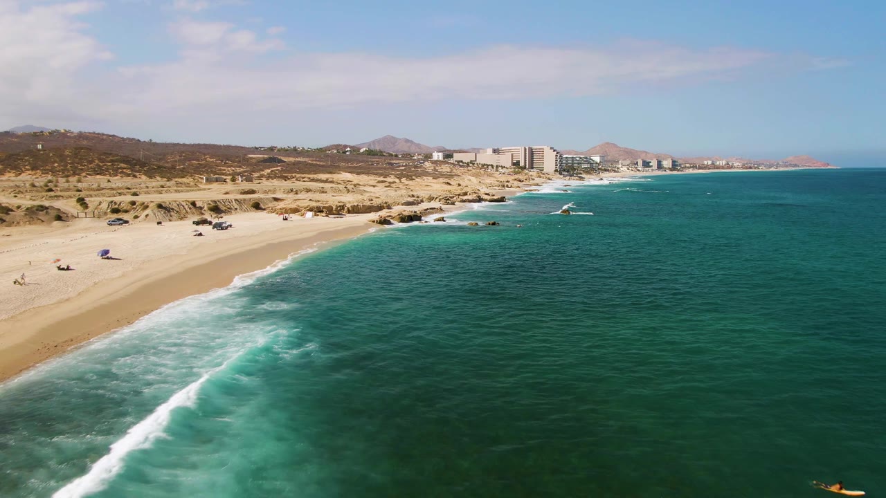 Aerial view at El Tule Beach in Los Cabos, Mexico, with turquoise water, white surf, a broad sandy beach, scrub hills, distant buildings, and tiny beach activity near the shore.