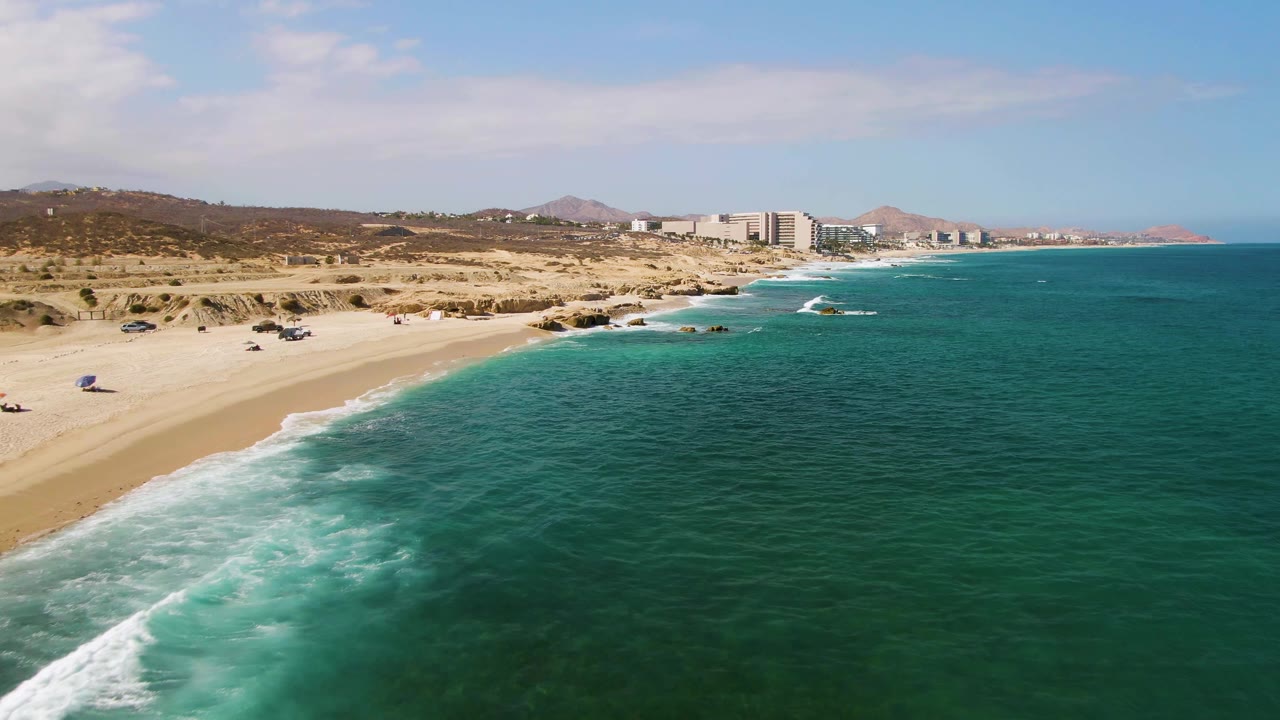 Wide coastal aerial with waves washing onto sand and resort buildings beyond the beach.