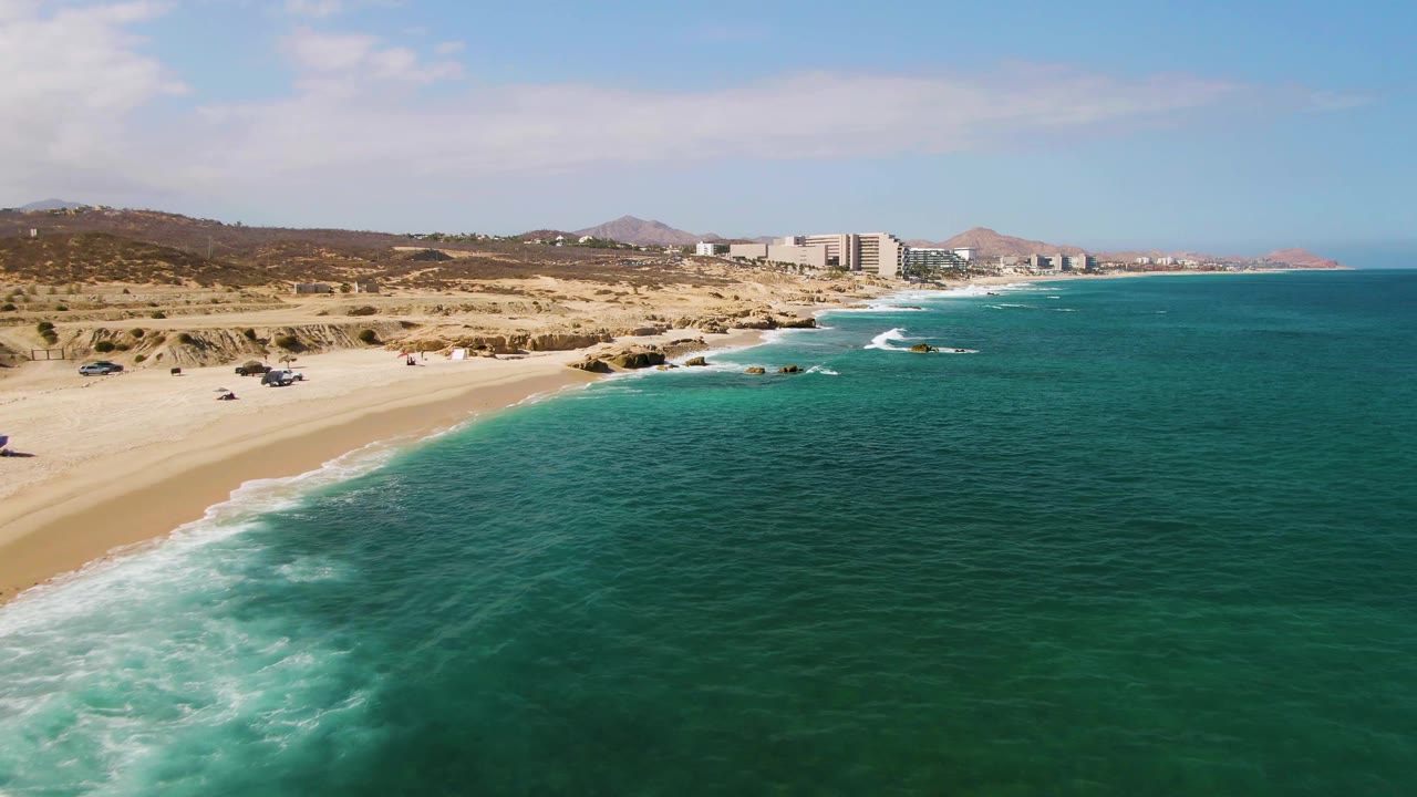 Aerial frame over El Tule Beach with foamy surf reaching the sand, several small vehicles or beach setups on the left beach, distant buildings near the center, and open water dominating the right side.