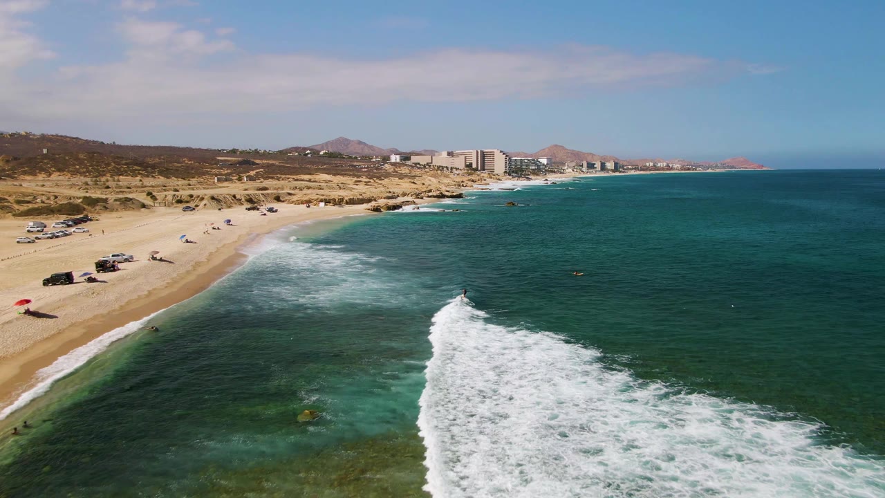 At El Tule Beach in Los Cabos, a white wave breaks diagonally toward the sandy shore while parked cars, small umbrellas, distant buildings, and small visible people line the beach.