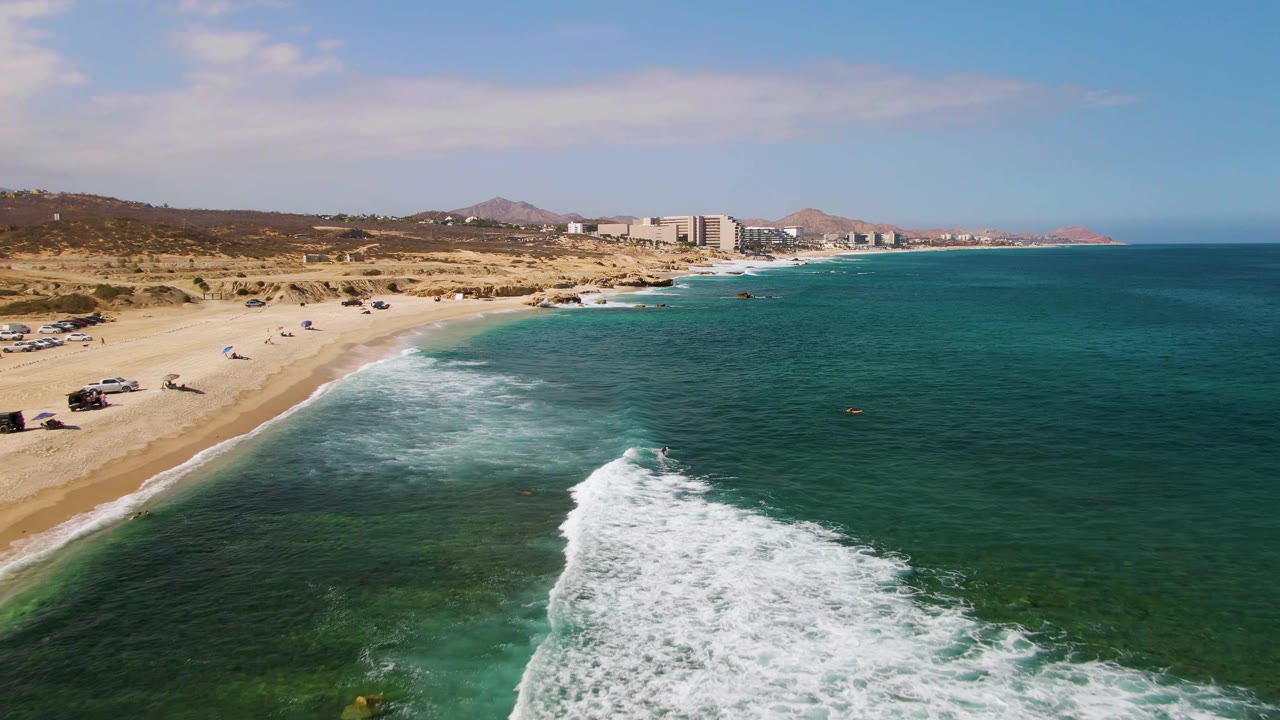 Drone view of surf rolling toward a sandy shoreline with parked beach vehicles.