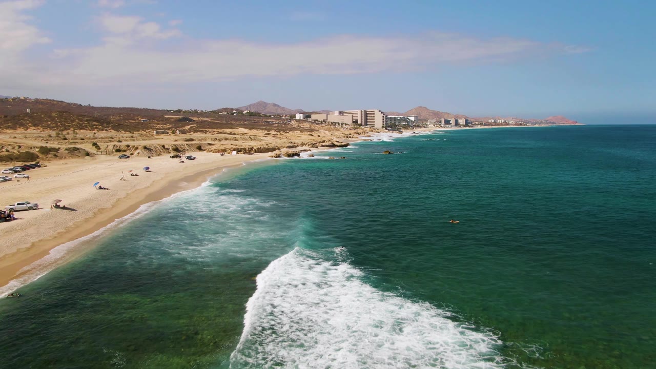 At El Tule Beach in Los Cabos, the foamy wave sits farther right in the frame, with clear turquoise water over the nearshore rocks or sand, a wide beach at left, parked cars, and tiny visible people.