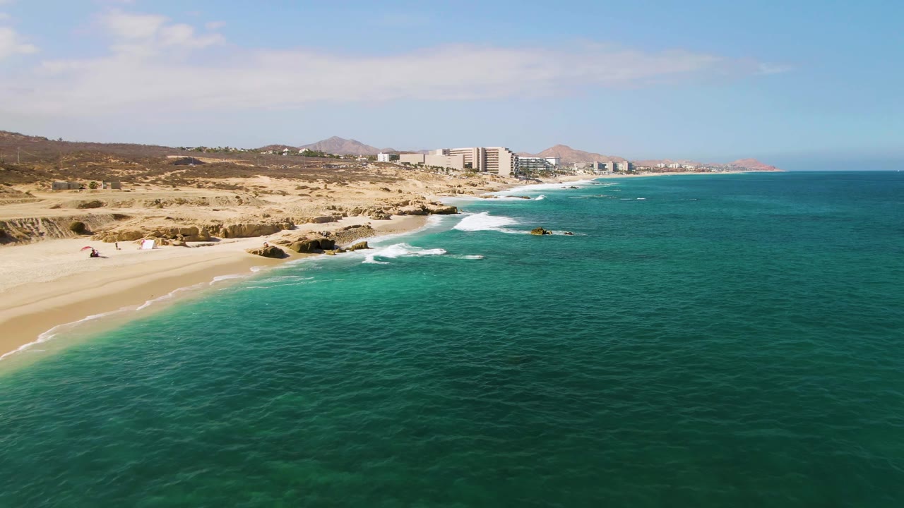 Aerial view over El Tule Beach in Los Cabos, with turquoise water in the foreground, sandy beach at left, rocky surf along the shore, distant buildings and hills, and no visible people.
