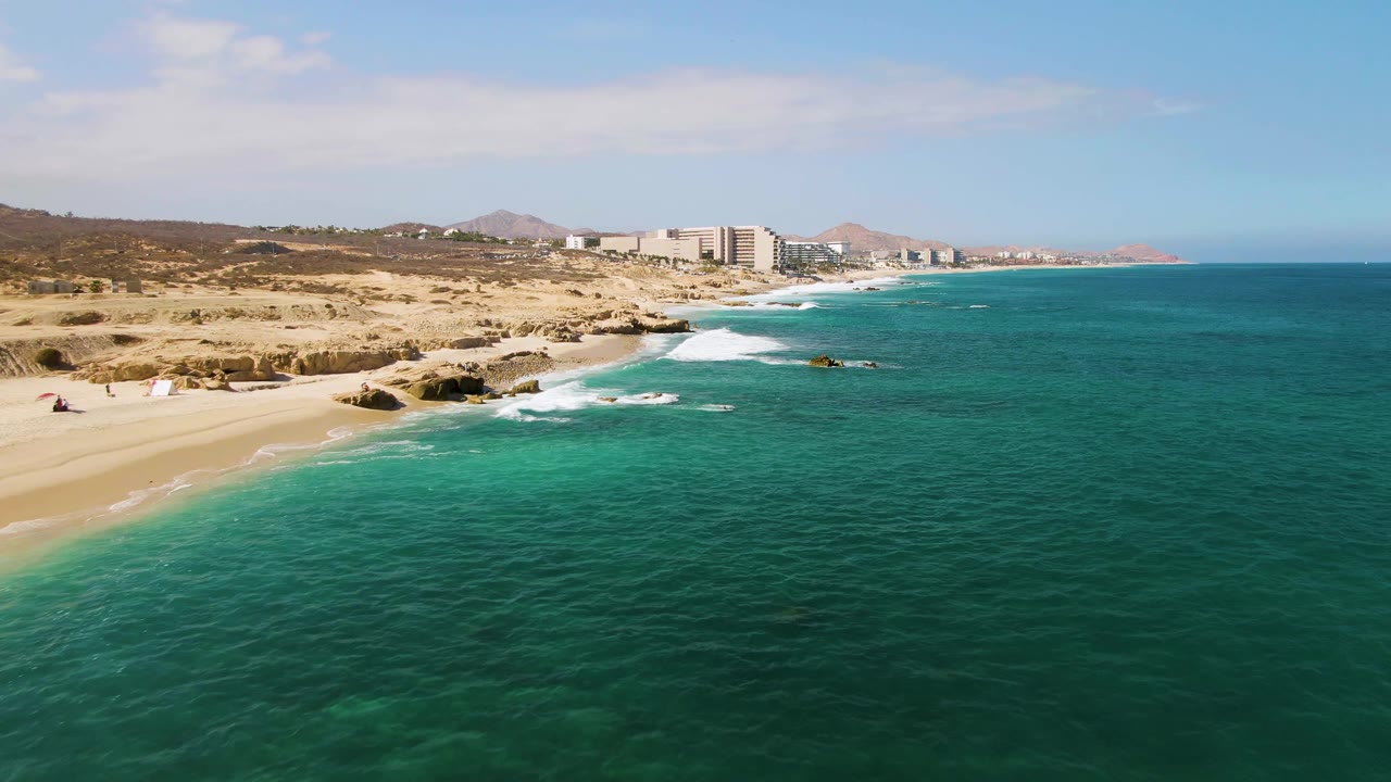 Turquoise water and white surf along rocks near a sandy Los Cabos beach.