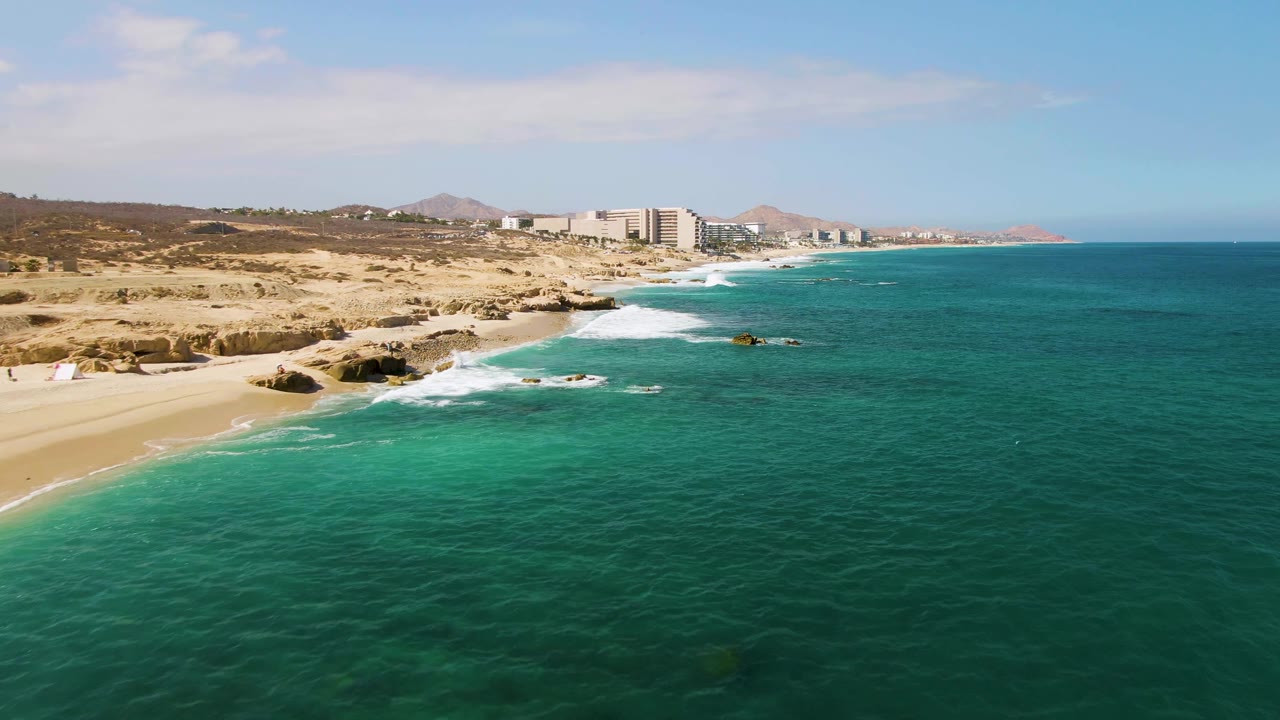 Aerial view of El Tule Beach with the shoreline running diagonally through frame, waves breaking over dark rocks, clear green-blue water across the foreground, distant buildings, and no visible people.