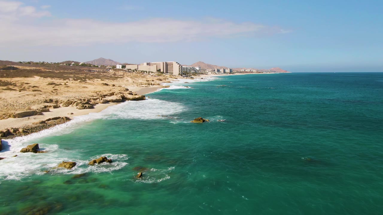 Drone view at El Tule Beach Los Cabos with foamy waves spreading along the sandy and rocky shore, resort-like buildings in the distance, and no visible people.
