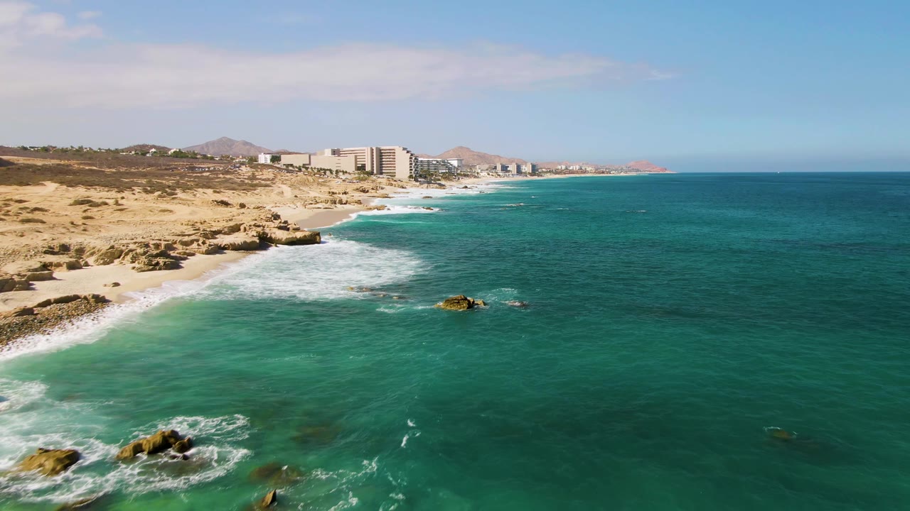 High coastal view of El Tule Beach Los Cabos where white breakers trace the shore, small rocks sit in the surf, beachfront buildings line the background, and no visible people appear.