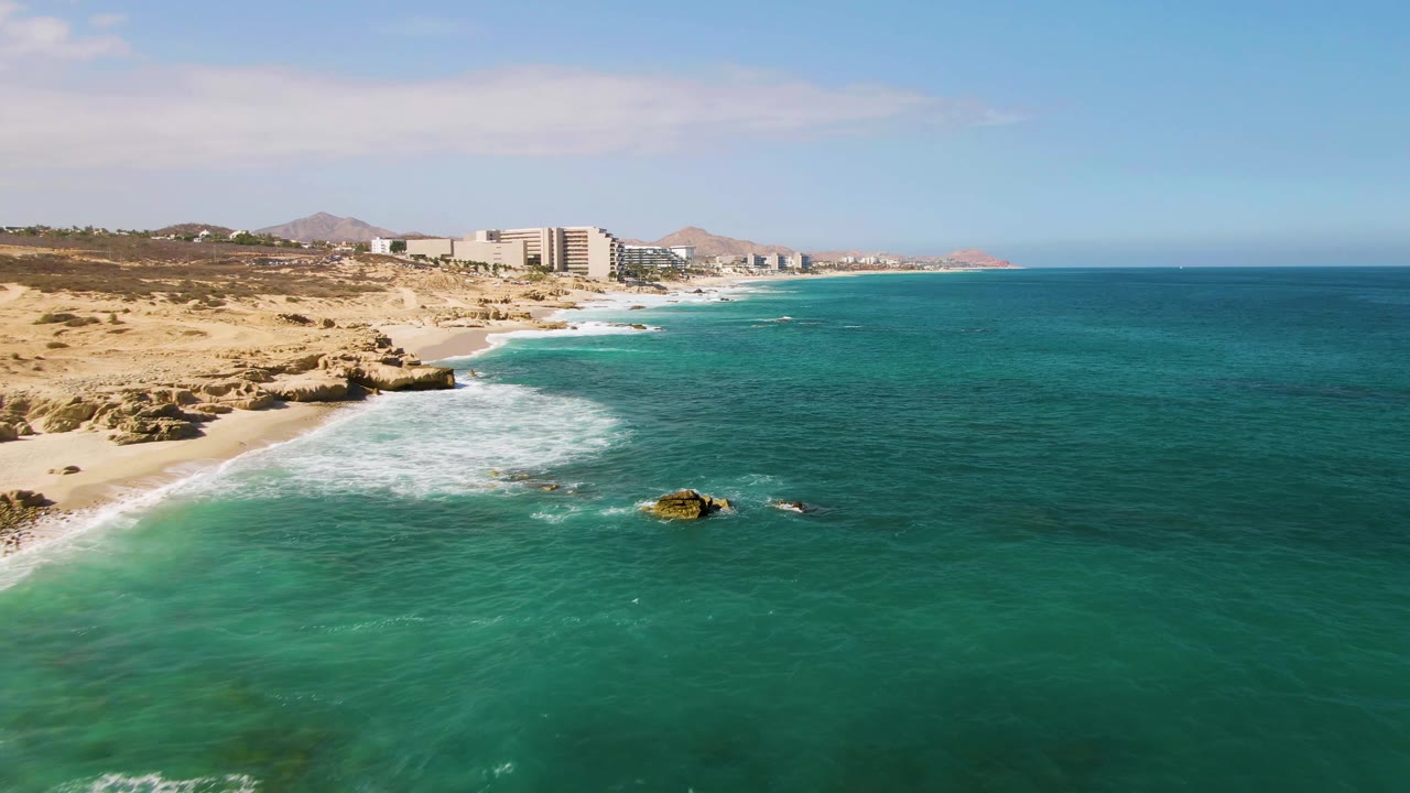 Aerial frame from El Tule Beach Los Cabos showing white surf wrapping around a rock near the beach, turquoise water in the foreground, distant buildings, and no visible people.