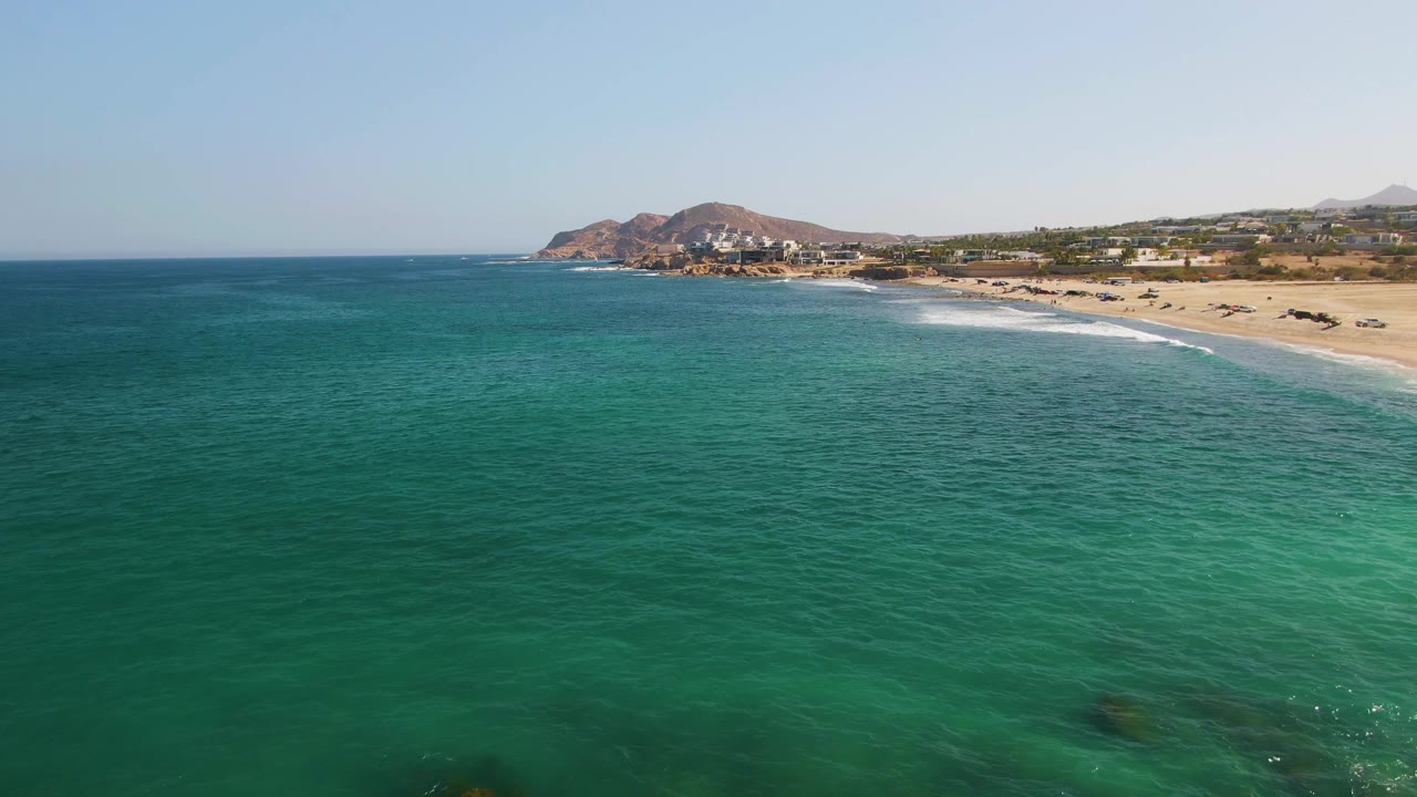 Aerial view of turquoise ocean beside a sandy beach and low hills.