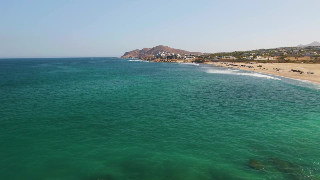 Aerial frame at 0:01 over green-blue ocean at El Tule Beach, with a thin line of white surf reaching the sand, hillside buildings in the distance, and no visible people.