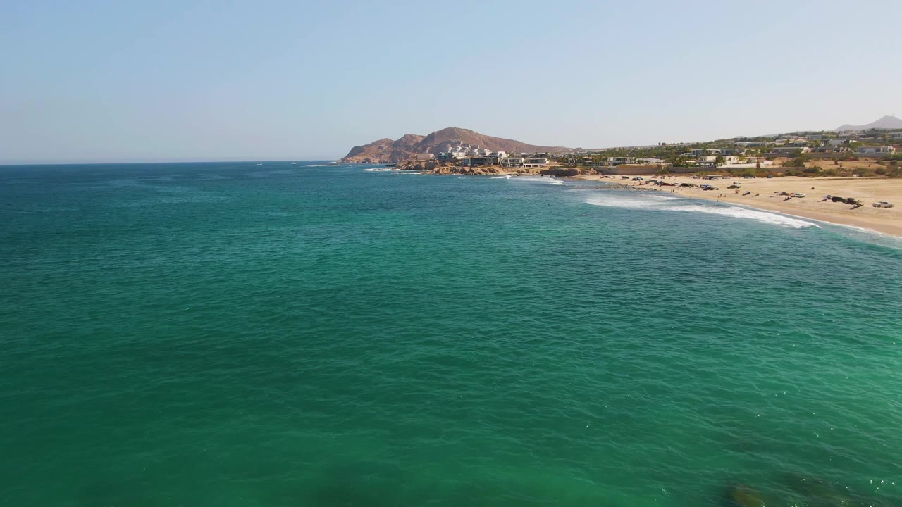 Aerial view at 0:02 of clear turquoise water off El Tule Beach, with the beach angled across the right side, low surf, distant hills, shoreline buildings, and no visible people.