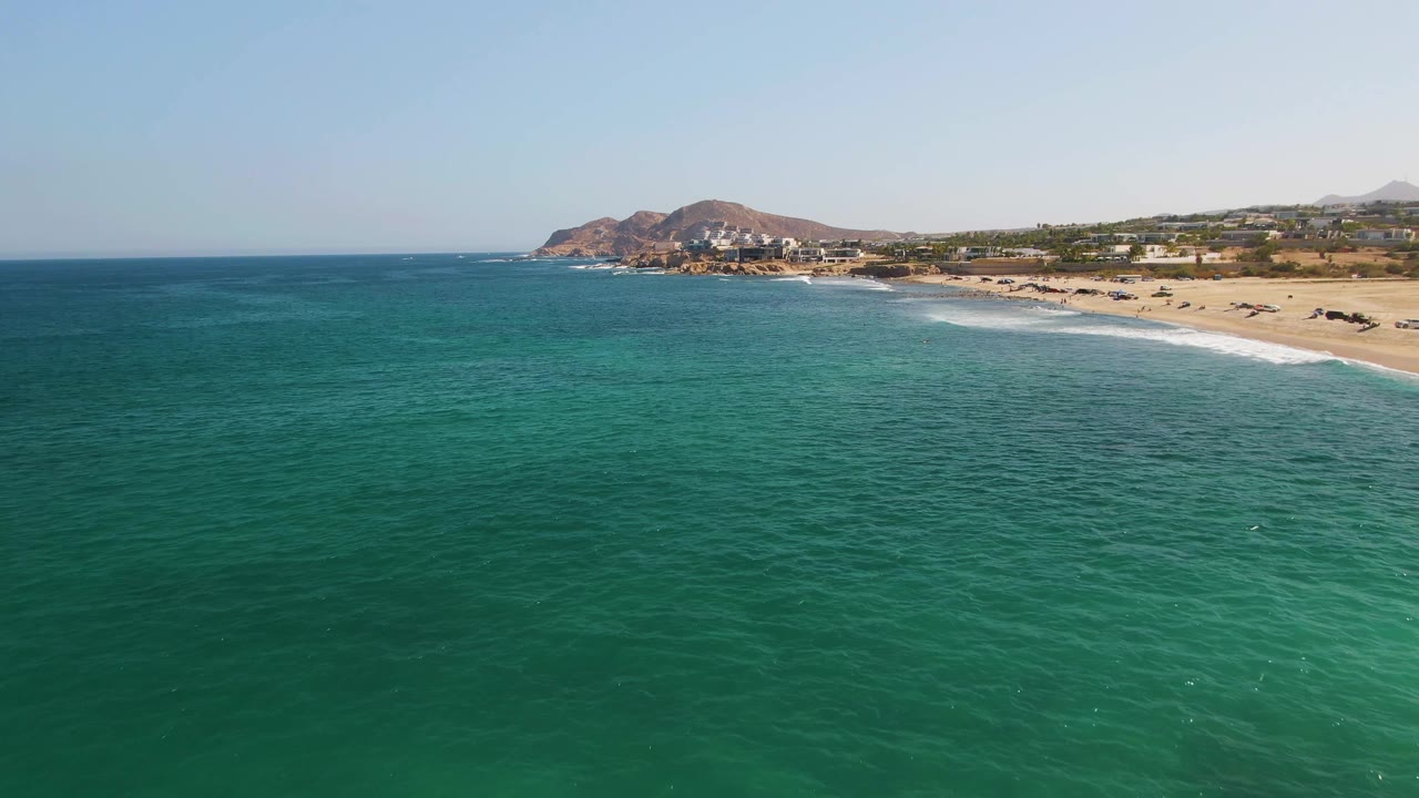 At 0:03, the aerial composition keeps the ocean foreground broad, with gentle waves rolling toward El Tule Beach, hills and small buildings behind the sand, and no visible people.