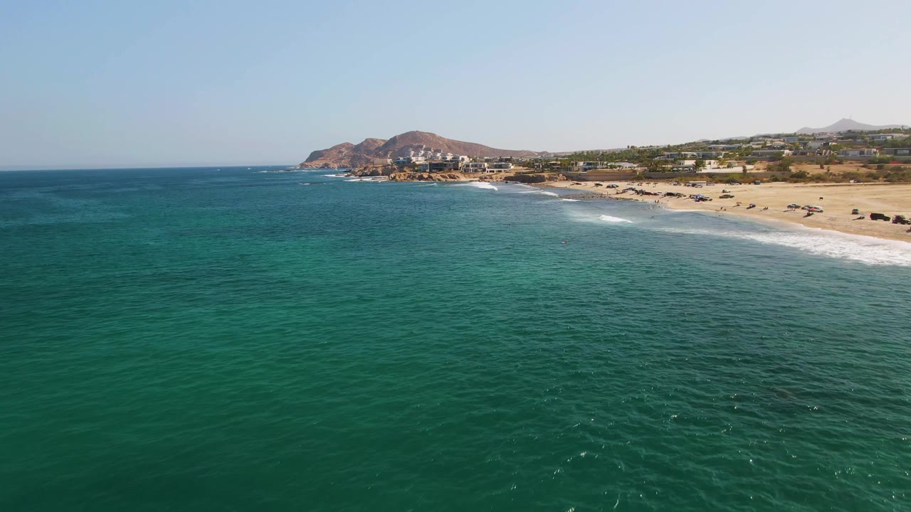 Drone view over El Tule Beach in Los Cabos with calm turquoise ocean filling most of the frame, a narrow surf line along the sand, hillside terrain in the distance, and no visible people.