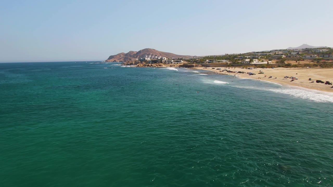 Aerial view of El Tule Beach in Los Cabos showing textured turquoise sea, small white waves approaching the sandy beach, low coastal buildings, brown hills, and no visible people.