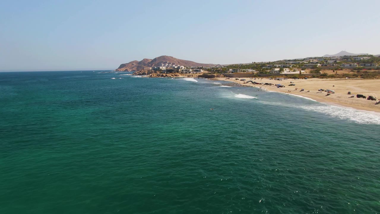 Aerial frame over El Tule Beach in Los Cabos with green-blue water in front, the sandy beach running diagonally along the right side, rocky hills beyond the shoreline, and no visible people.