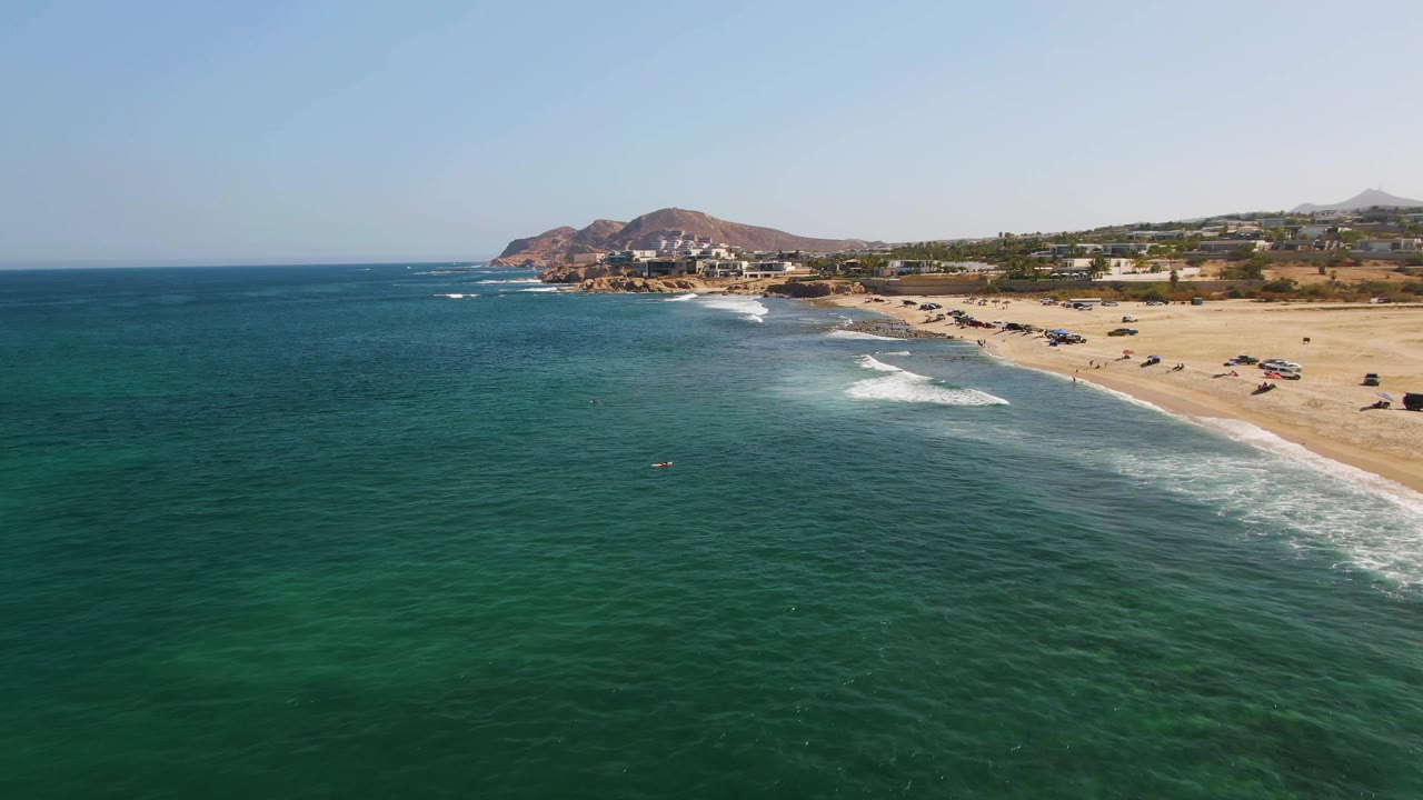 Aerial view at El Tule Beach in Los Cabos with green-blue water in the foreground, white surf approaching the sand, distant buildings and hills, and no visible people.