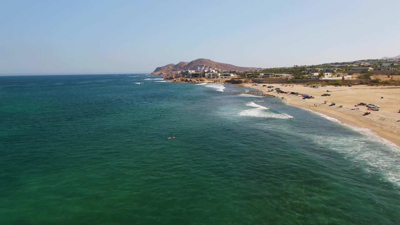 Drone view of surf bands rolling toward a sandy beach with buildings beyond.