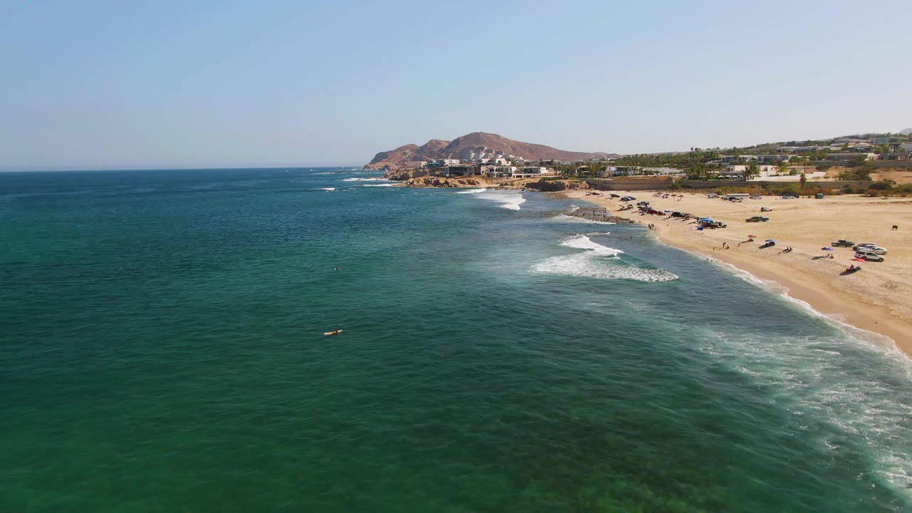 Wide aerial frame from El Tule Beach in Los Cabos showing darker green water in front, a white wave breaking near shore, a sandy beach with parked vehicles, distant buildings, and no visible people.