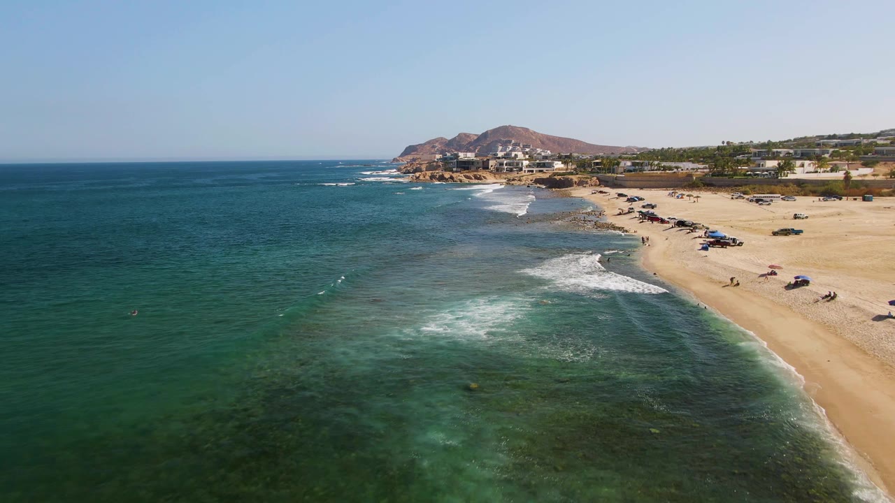 Aerial view at El Tule Beach in Los Cabos showing turquoise water in the foreground, a sandy shoreline to the right, umbrellas on the beach, and low buildings below reddish hills in the distance.