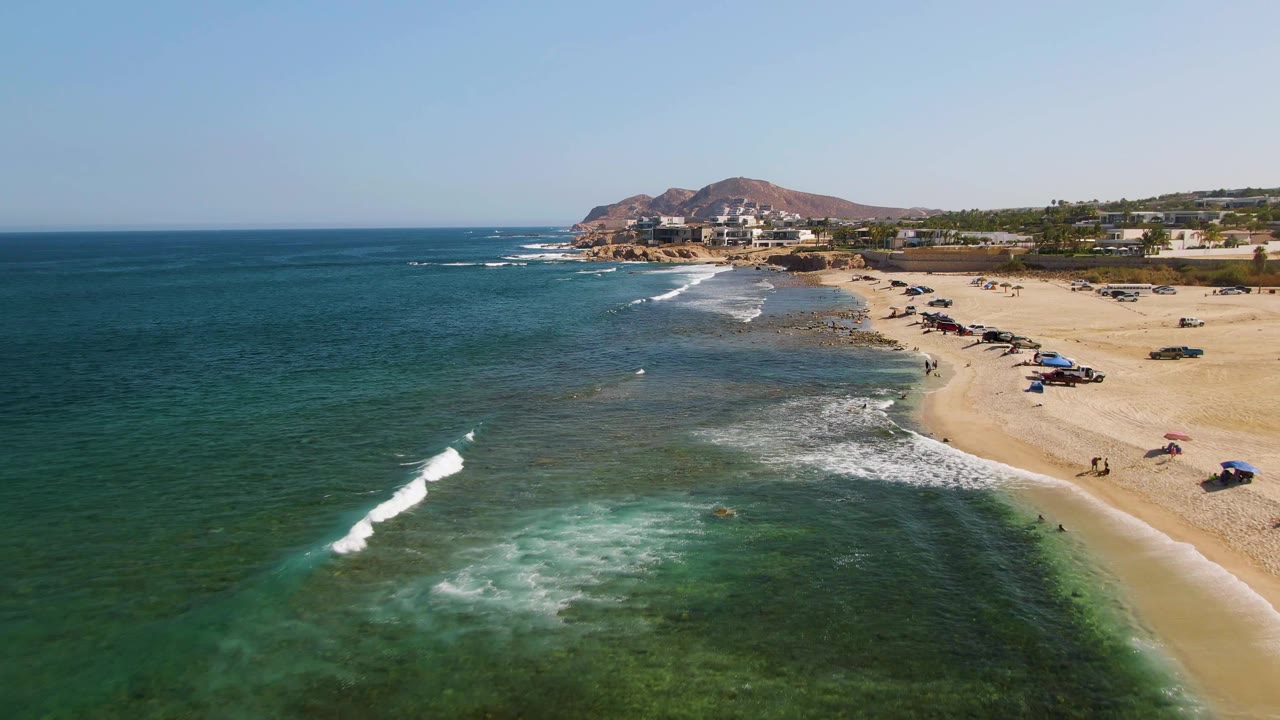 At 0:02, a narrow white wave line advances through green-blue water toward El Tule Beach, with umbrellas and small beachgoers on the sand and buildings and hills beyond the shoreline.