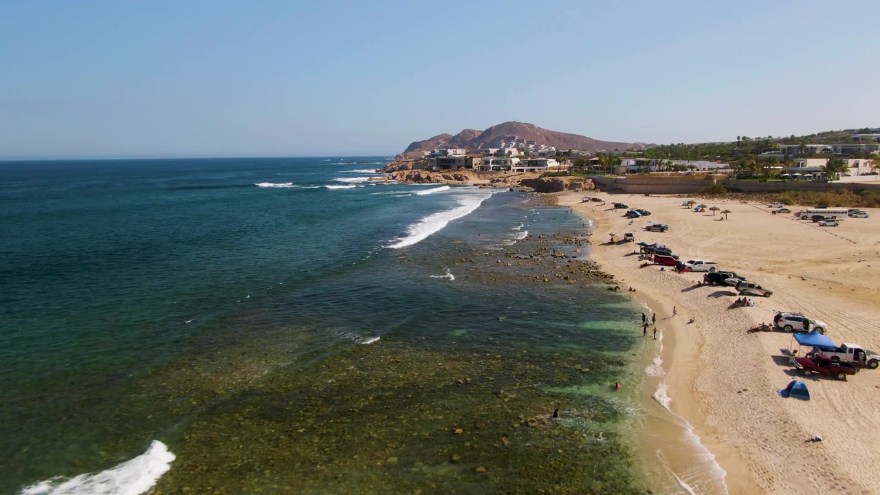 Aerial frame of El Tule Beach in Los Cabos showing dark blue ocean, green shallows, white surf, parked vehicles on pale sand, small visible beachgoers, low buildings, and hills beyond the shoreline.